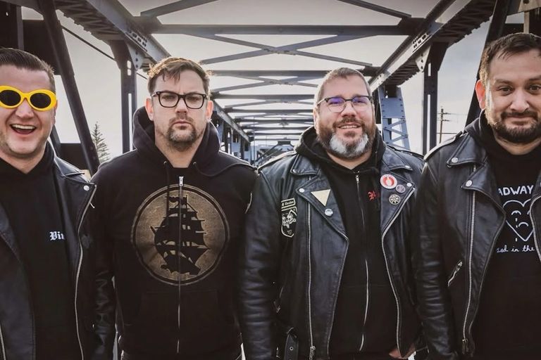 Punk rock band Teenage Bottlerocket posing together outdoors on a steel truss bridge in black leather jackets, hoodies, and yellow sunglasses.